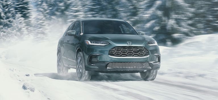 2027 Honda HR-V EX-L in Nordic Forest Pearl front driving through a snowy winter landscape (Subcompact Crossover SUV).